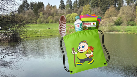 Im Hintergrund ein See umgeben von Wiesen. Davor ein grüner Stoffbeutel, aus dem eine Wasserflasche, eine Feder, Papiere und ein Heft hinausschauen.