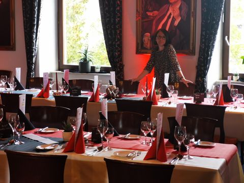 L'image montre une salle à manger élégamment décorée, probablement préparée pour un événement ou une célébration formelle. L'accent est mis sur plusieurs longues tables, recouvertes de nappes blanches et de chemins de table rouges. Chaque place comprend des couverts polis – fourchettes à droite des assiettes, couteaux derrière et cuillères complètement à gauche. Des serviettes rouges pliées en cônes triangulaires servent de centre de table sur chaque assiette.