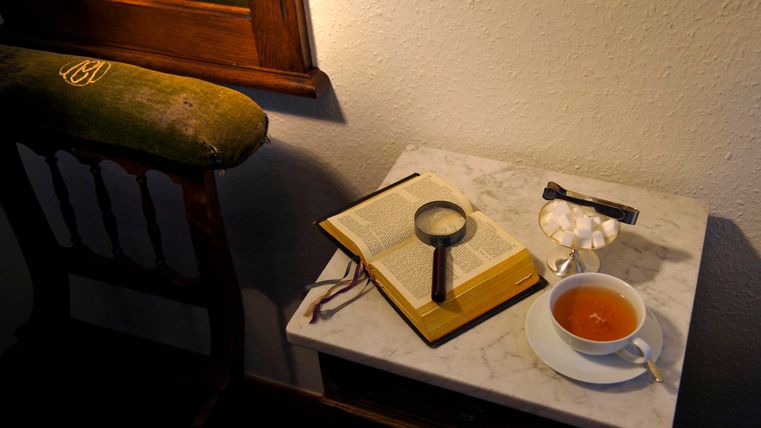 Une table avec un livre, une loupe et une tasse de thé. En arrière-plan, on peut voir un miroir et une chaise.