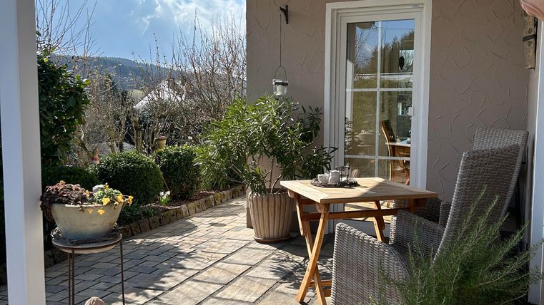 Un espace extérieur accueillant avec une table en bois et des chaises. Entouré de plantes et d'un ciel bleu clair.