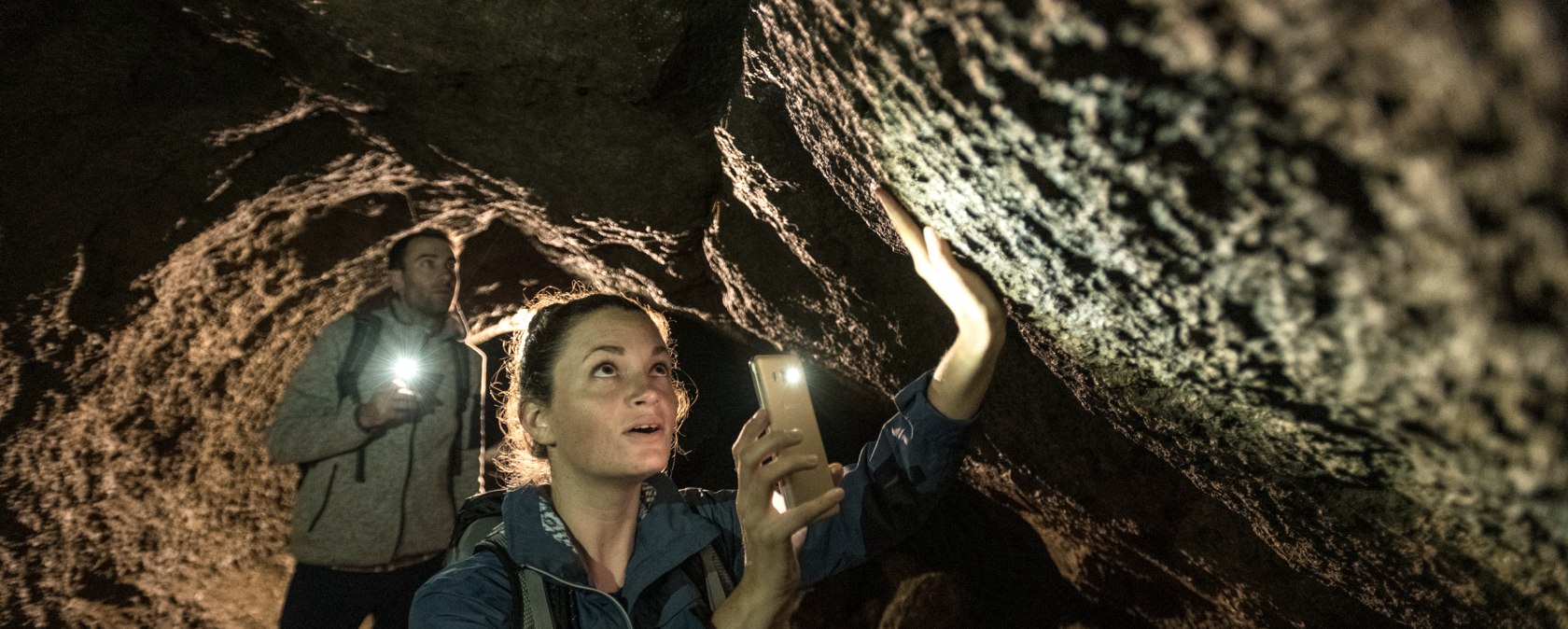 Two people in a dark cave shine the torches of their mobile phones along the rock faces.