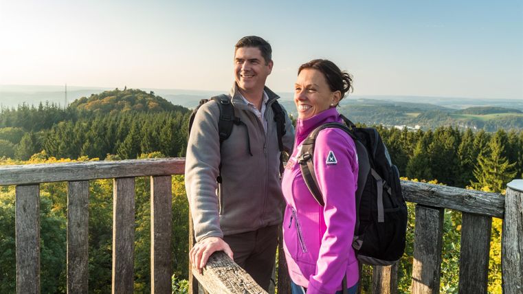 Ein Mann und eine Frau stehen lächelnd auf einem Aussichtsturm, umgeben von Wald und Hügeln im Hintergrund.