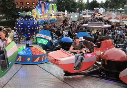 Fahrgesch&auml;ft "BreakDancer" mit Passagieren. Dahinter buntes Kirmestreiben mit vielen Besuchern und Schaustellern.