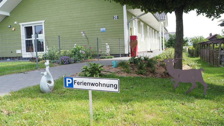 Ein Ferienhaus mit einem Parkplatzschild und einer hübschen Grünfläche. Im Vordergrund sind dekorative Elemente wie eine Statue und ein stilisiertes Tier zu sehen.