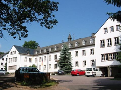 Blick auf eine großes dreistöckiges Gebäude in L-Form. Auf dem schwarzgedecktes Dach sind elf Dachgauben angebracht. Hinder dem Gebäude schaut die Spitze eines  Glockenturms hervor.
Vor dem Gebäude ist ein Parkplatz mit mehreren Autos.