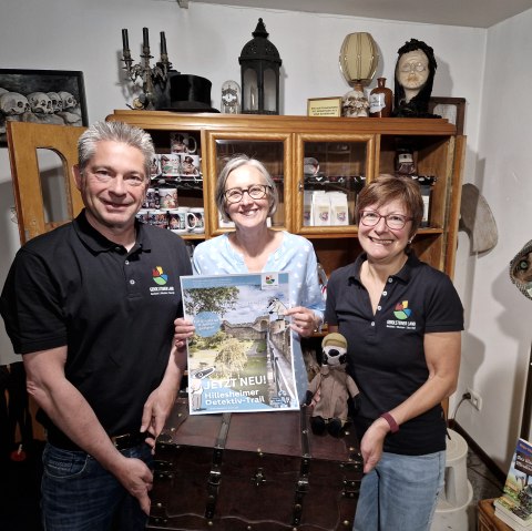 Drei l&auml;chelnde Personen stehen in einem kleinen Raum vor einem Holzschrank und halten eine gro&szlig;e Schatzkiste und ein Plakat zum Detektiv-Trail hoch.