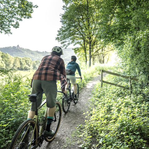 Zwei Radfahrer auf Gravelbikes fahren auf einem schmalen Naturpfad neben einem dichten Wald.