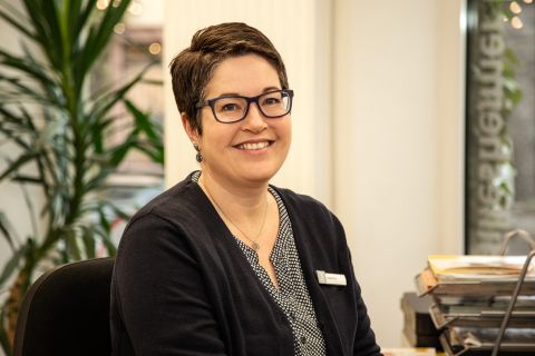 Eine Frau sitzt an einem Schreibtisch und lächelt in die Kamera. Hinter ihr ist eine große Palmenpflanze.