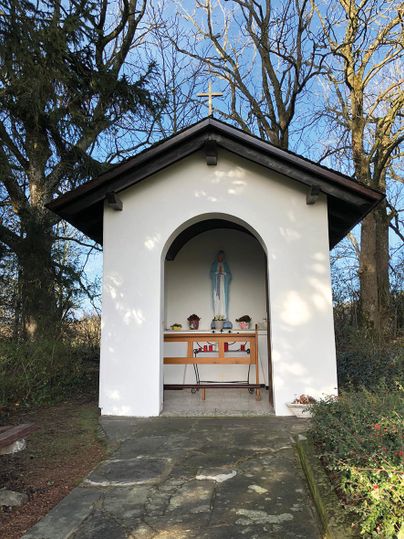Kleine weiße Kapelle mit Marienstatue und Kreuz auf dem Dach, umgeben von Bäumen.