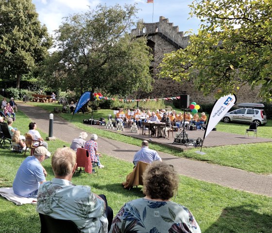 zahlreiche Personen sitzen auf Stühlen und Decken auf einer Wiese im Park und schauen zur kleinen Bühne mit Musikverein und Lesern vor der Hillesheimer Stadtmauer