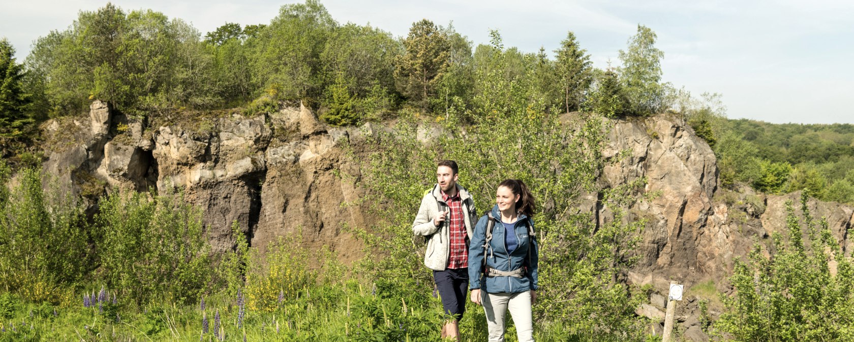 Two people in hiking gear walk along a path through the Steffeln Volcano Garden, surrounded by green vegetation and rocks.