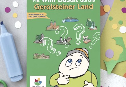 Titelseite  des R&auml;tselhefts "Mit Willi Basalt durch das Gerolsteiner Land" auf einem Tisch voller Mal- und Bastelutensilien und bunter Bl&auml;tter.