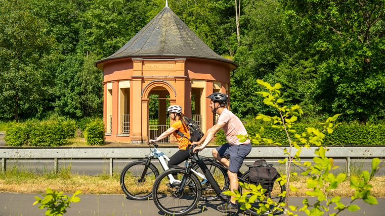 Zwei Personen mit Fahrrädern stehen auf einer Brücke vor einem achteckigen Pavillon aus rotem Stein, umgeben von grünen Bäumen.