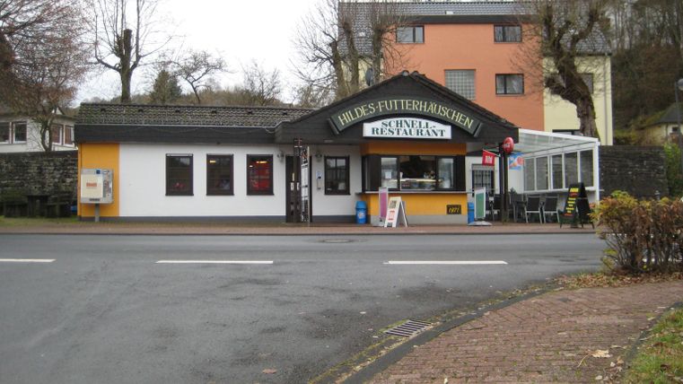 Hildes Futterhäuschen in Stadtkyll, ein Schnellrestaurant mit gelber und weißer Fassade, umgeben von kahlen Bäumen und Wohnhäusern.