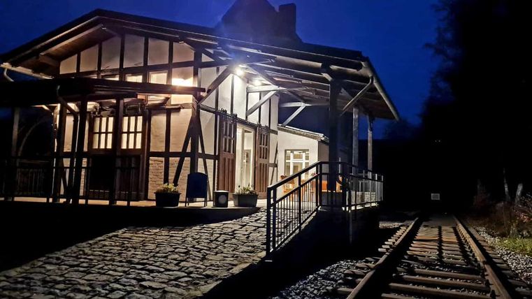 Ein historischer Bahnhof bei Nacht, beleuchtet von warmem Licht. Der Weg zu den Gleisen ist durch Steine und eine Rampe gekennzeichnet.