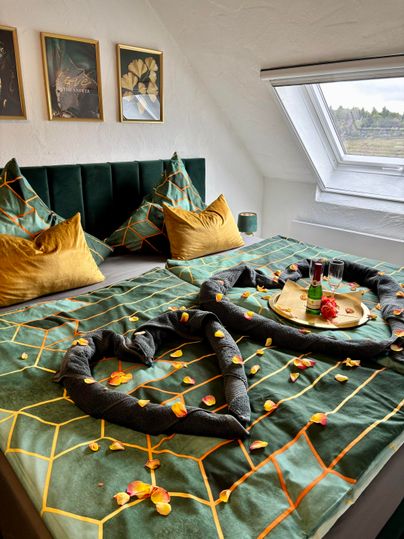 Ein elegantes Schlafzimmer mit einem großen Bett, das mit grüner Bettwäsche und goldenen Kissen dekoriert ist. Auf dem Bett liegt eine romantische Anordnung aus Handtüchern und Blütenblättern, daneben steht ein Tablett mit Getränken und Snacks.