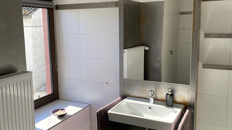 Ein modernes Badezimmer mit hellen Fliesen und einem großen Spiegel. Die Ausstattung umfasst ein Waschbecken, eine Heizkörper und ein Fenster für Tageslicht.