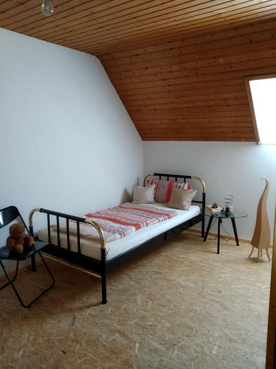Une chambre simple avec un lit simple et une chaise. Les murs sont blancs et le plafond est en bois, les fenêtres laissent entrer beaucoup de lumière.
