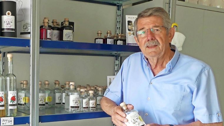 Ein älterer Mann steht vor einem Regal mit verschiedenen Flaschen. Er hält eine Flasche in der Hand und schaut freundlich in die Kamera.