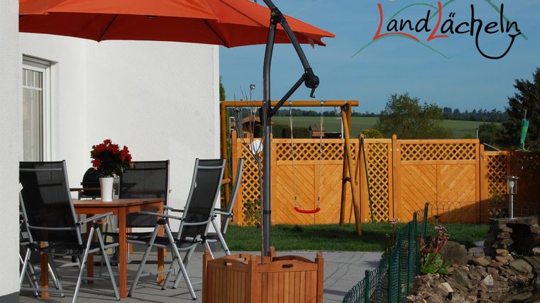 Ein gemütlicher Außenbereich mit einem Tisch und Stühlen unter einem orangefarbenen Sonnenschirm. Im Hintergrund sind ein Spielplatz und eine Holzlattenzaunlandschaft sichtbar.