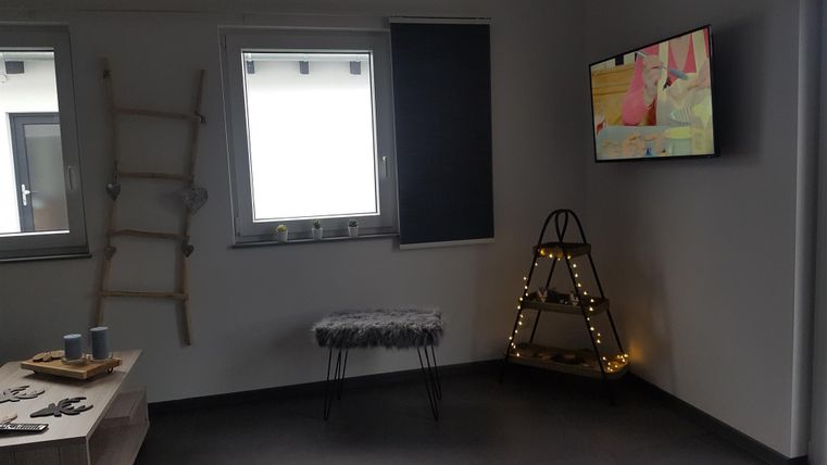 Ein moderner Raum mit einem Fernseher an der Wand. Zu sehen sind Fenster, ein Hocker und eine dekorative Leiter.