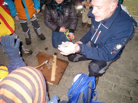 Ein Mann zeigt einer Gruppe von Kindern einen Stein. Sie stehen auf einem gepflasterten Boden, umgeben von Outdoor-Kleidung.