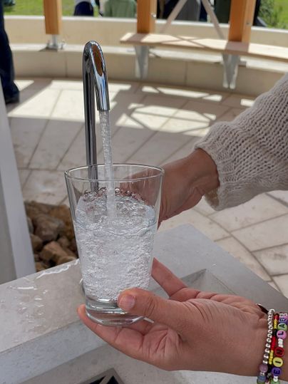 Een glas wordt gevuld met bruisend water uit een kraan; op de achtergrond zijn mensen en een paviljoen te zien.