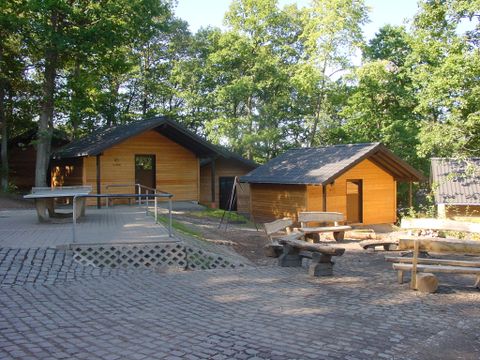 Holzhütten in einem Wald, umgeben von Bäumen. Im Vordergrund sind Sitzbänke und eine Tischtennisplatte zu sehen.