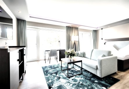 Modern eingerichtetes Hotelzimmer mit einem gro&szlig;en Doppelbett,  davor eine Couch auf einem Teppich mit Tisch und einem gro&szlig;en Flachbildfernseher an der Wand. Vor der gro&szlig;en Fensterfront steht ein Tisch mit einem Stuhl.