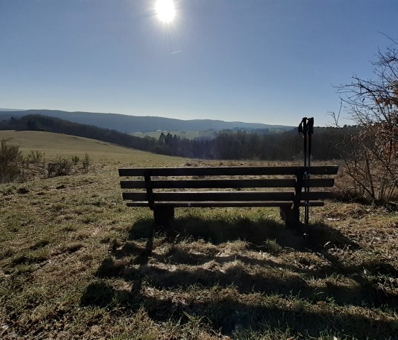 Bank auf einem Feld mit Wanderstöcken, Blick auf Hügel und strahlende Sonne am Himmel., © Touristik GmbH Gerolsteiner Land, Ute Klinkhammer