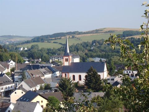 Blick von oben auf den Ort Stadtkyll, in deren Mitte die Kirche St. Josef steht.Das Gebäude ist im klassizistischen Stil errichtet,mit einem einfachen viereckigen Mittelschiff. Rechts ist der Glockenturm mit einem hohen, spitzen,schwarz eingedecktem Dach. Das Gebäude ist in weiß gestrichen, die Rundfenster sind, genauso wir die Ecken des Glockenturm ,rötlich abgesetzt.