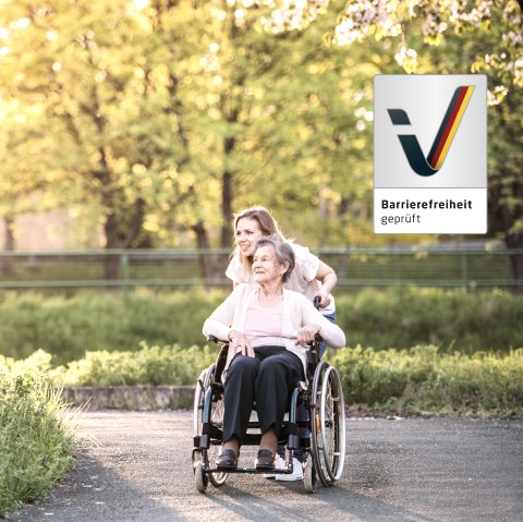 An elderly lady in a wheelchair is pushed along a path surrounded by meadows by a younger woman. The two are in a good mood. Next to them is the &lsquo;Accessibility tested&rsquo; logo.
