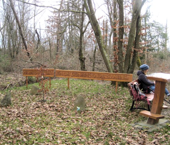 A person is sitting on a bench in the forest. A sign with the inscription 'Scherre Kreuz, a place of peace and reflection' is visible., © Touristik GmbH Gerolsteiner Land, Ute Klinkhammer