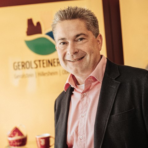 A smartly dressed gentleman in a shirt and jacket smiles at the camera. Behind him is the logo of Touristik GmbH Gerolsteiner Land and some souvenir items.