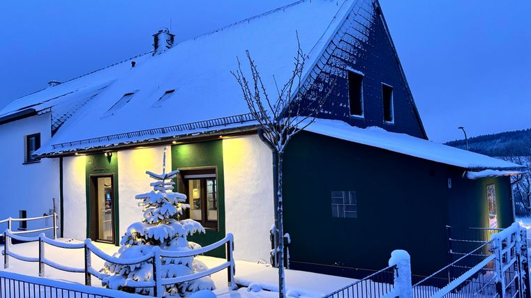 Ein gemütliches Haus im Schnee mit einem beleuchteten Eingangsbereich. Ein kleiner, geschmückter Baum steht vor dem Gebäude.