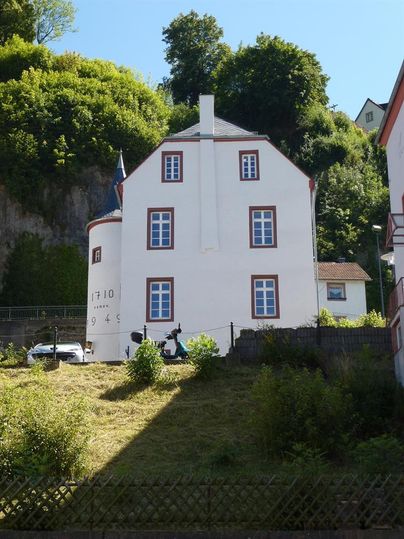 Weißes Gebäude mit roten Fensterrahmen, umgeben von grünen Bäumen und einem Hang. Ein Auto und eine Skulptur sind im Vordergrund sichtbar.