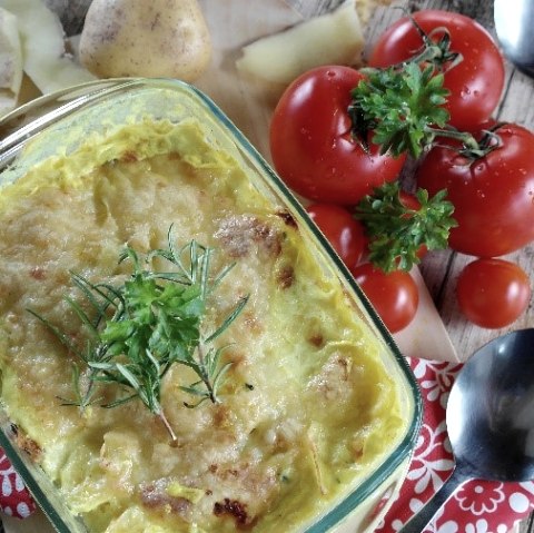 A glass baking dish with a cheese-topped potato dish in it. Next to it are tomatoes, potatoes and spoons.