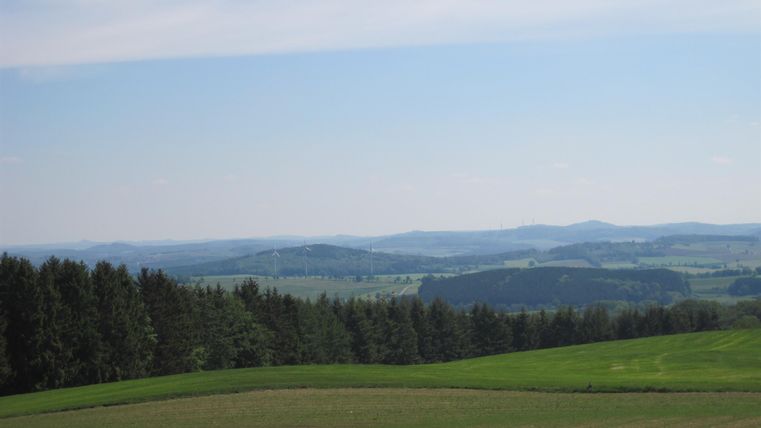 Blick über grüne Felder und Wälder in der Eifel, mit Windrädern am Horizont und blauem Himmel.