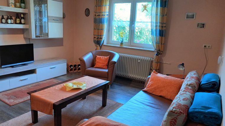 A cozy living room with a sofa, an armchair, and a coffee table. A TV and shelves with bottles are also available.