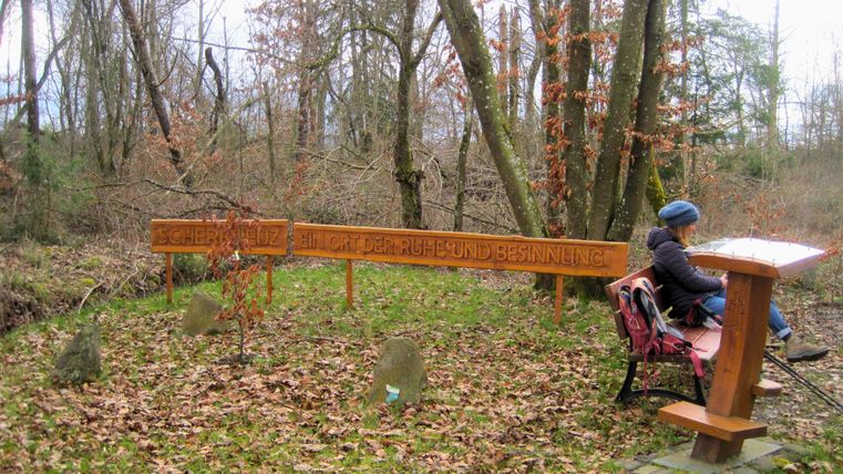 Een persoon zit op een bankje in het bos naast een bord met de tekst 'Scherre Kreuz, een plaats van rust en bezinning'.