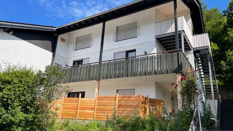 Une maison moderne avec un balcon et un revêtement en bois à l'avant. Entourée de plantes vertes et d'un ciel bleu.