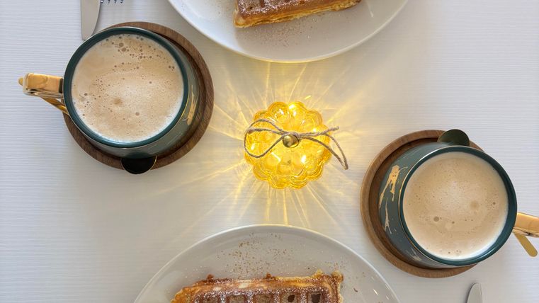 Zwei Teller mit Waffeln, jeweils garniert mit Sahne und Früchten, stehen auf einem Tisch. Dazu gibt es zwei Tassen Bier und eine kleine Lichtdekoration in der Mitte.