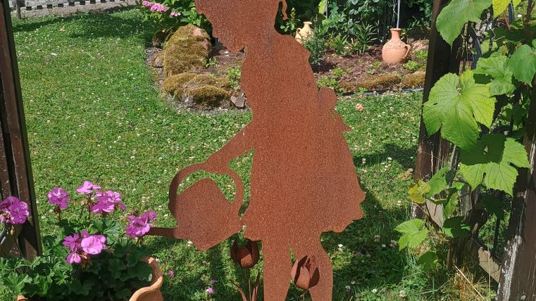 Eine rostige Metallfigur eines Mädchens, das mit einem Eimer in einem Garten steht. Im Hintergrund sind bunte Blumen und eine kleine Gartenbank zu sehen.