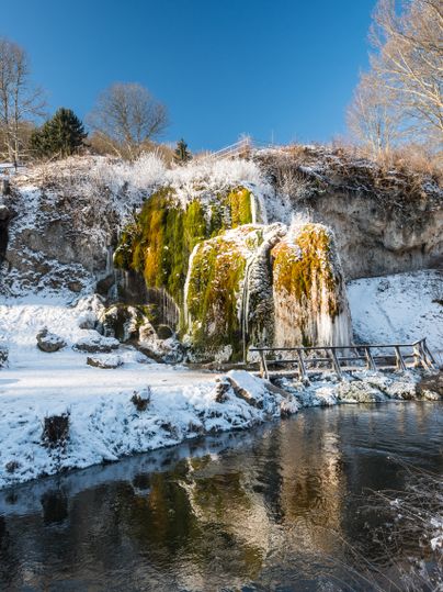 Verschneiter Wasserfall Dreimühlen mit Moos und Eis, umgeben von kahlen Bäumen und blauem Himmel.