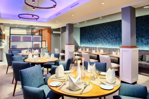 Modernes Restaurant mit blauen Stühlen, runden Holztischen und stilvoller Beleuchtung. Die Tische sind elegant gedeckt.