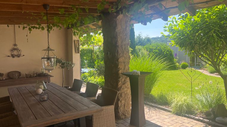 Un espace extérieur confortable avec une table en bois et des chaises. En arrière-plan, on peut voir des plantes vertes et un jardin bien entretenu.