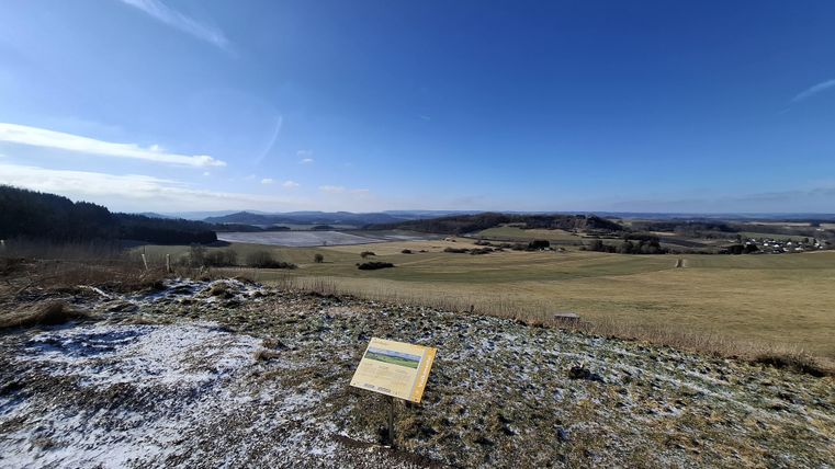 Eine weite Landschaft mit sanften Hügeln und klarem Himmel. Im Vordergrund steht ein Informationstafel.
