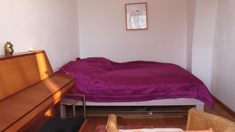 Een eenvoudige kamer met een bed in paarse beddengoed en een piano. Een ronde fauteuil en een schilderij aan de muur completeren de inrichting.