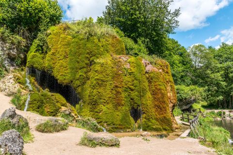 Moosbedeckter Wasserfall bei Nohn, umgeben von üppigem Grün. Ein kleiner Steg führt am Wasser entlang. Der Himmel ist blau mit einigen Wolken.