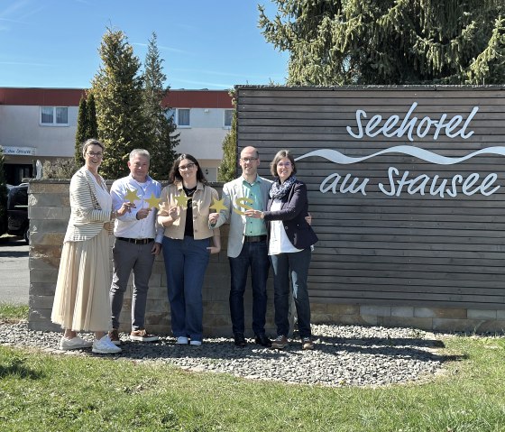 F&uuml;nf Personen stehen vor der Fassade des Seehotels am Stausee in gerolstein und halten jeweils 4 Sterne und ein "S" f&uuml;r die Sterneklassifizierung hoch.